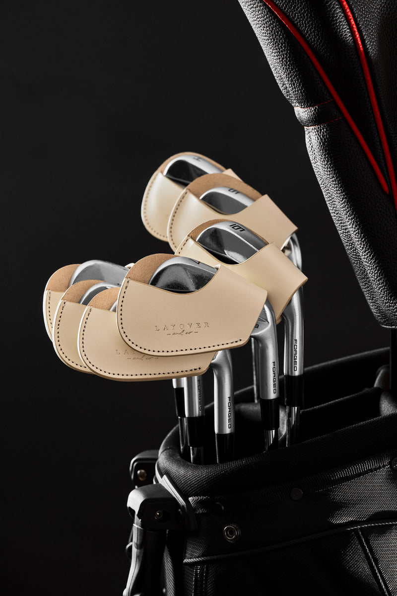 A pair of beige leather golf club covers resting on top of a black golf bag, with several metal golf clubs visible inside. The background is solid black.