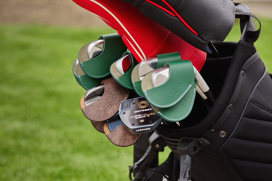Close-up of a golf bag with several golf clubs with iron covers, photographed outdoors on a blurred green grass background.