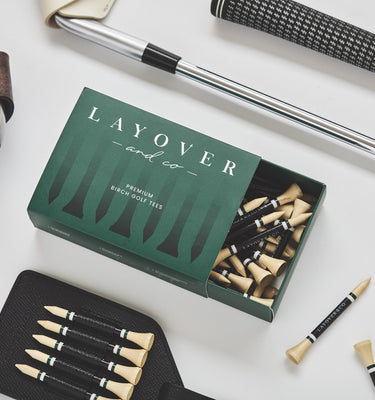A flat-lay product shot of a golf club accessory set on a white background, featuring a green box of Layover premium brass shop tees, black and gold clubbing tools, a leather strap, and metal shafts.