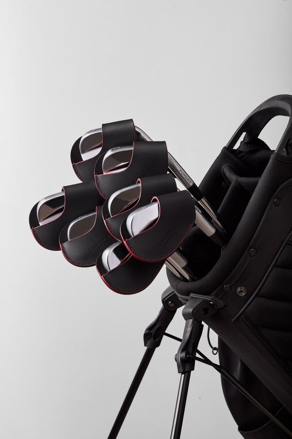 A close-up studio shot of a black golf bag with several black golf clubs inserted, featuring red trim accents and a light gray background.