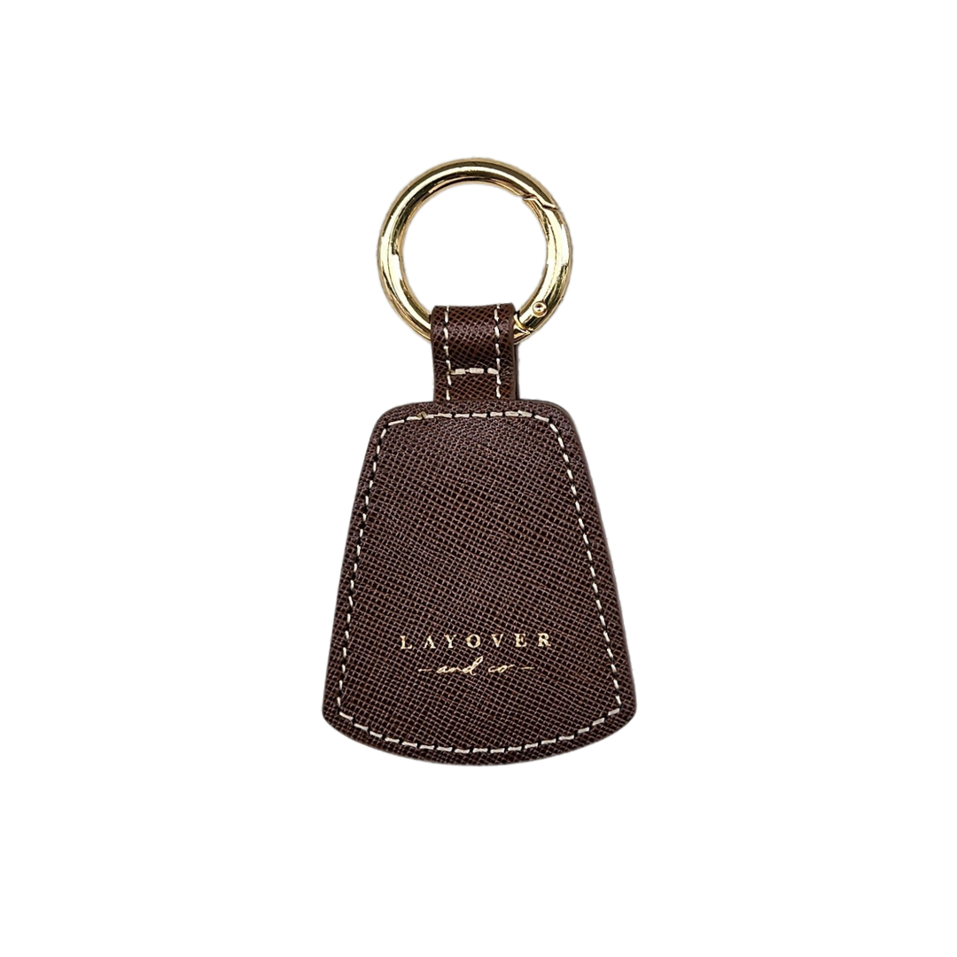 A premium brown leather hat holder from Layover & Co., featuring a textured finish, gold-embossed branding, and a sturdy gold keyring. Designed with a magnetic closure for securely holding hats on bags or belts.