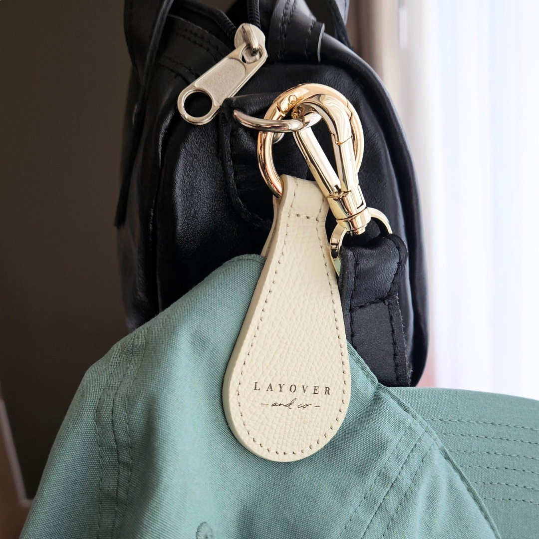 Cream-colored leather magnetic hat holder by Layover and Co. attached to a black bag with gold hardware, securely holding a green cap. Designed for stylish, hands-free convenience on the go.