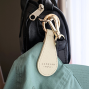 Cream-colored leather magnetic hat holder by Layover and Co. attached to a black bag with gold hardware, securely holding a green cap. Designed for stylish, hands-free convenience on the go.