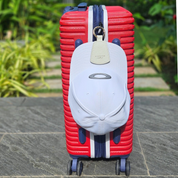 Red and navy hard-shell suitcase standing upright on a stone pathway, with a white leather hat holder from Layover and Co. attached to the handle. A white Oakley cap is secured by the hat holder, showcasing its stylish and practical design for travelers. The background features lush greenery, adding a vibrant contrast to the travel-ready setup.