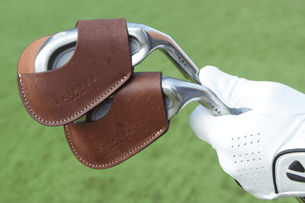 Golf club with a brown leather iron cover held by a gloved hand on a green background