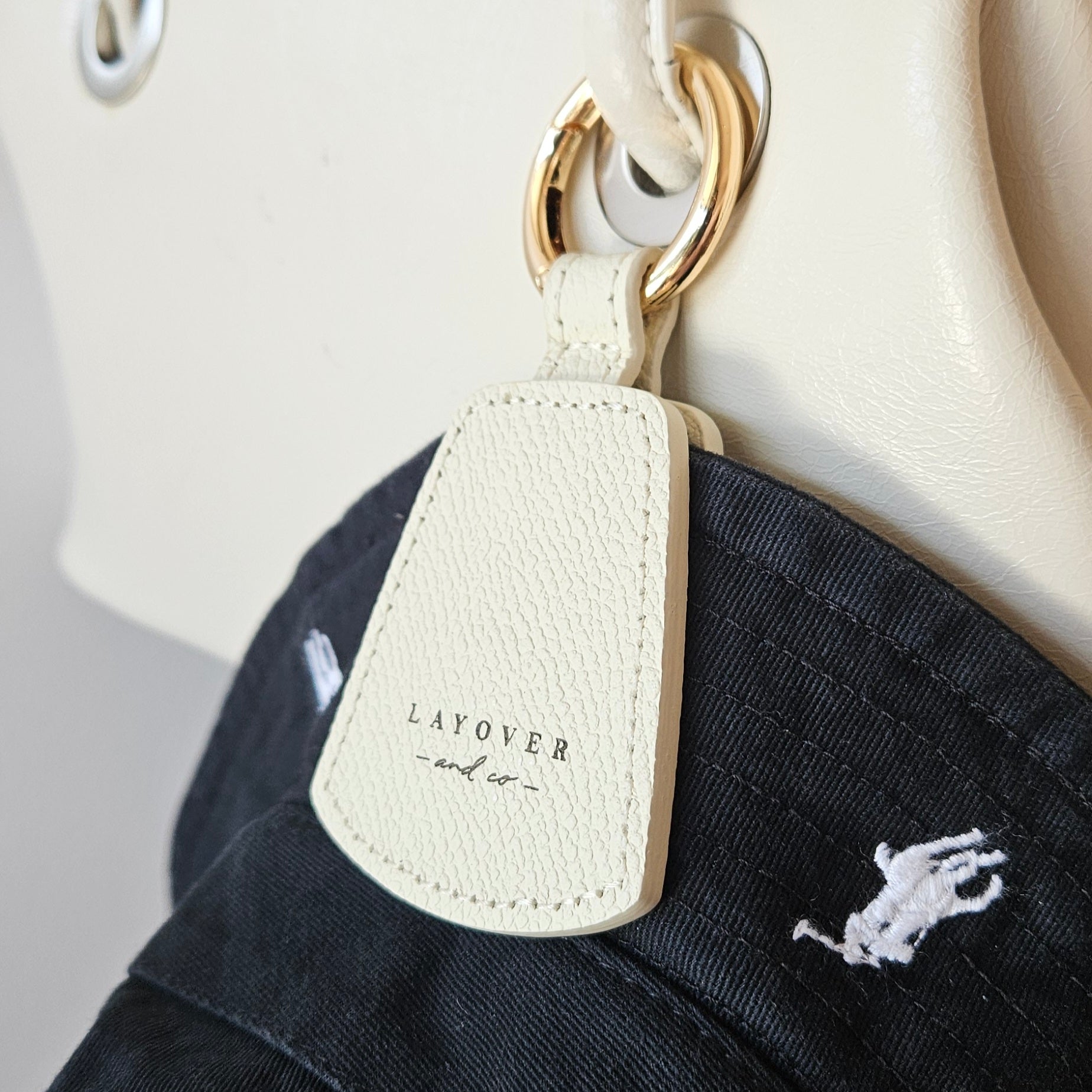 A close-up of Layover and Co. leather accessories, including a brown leather hat holder securing a white cap and a green leather sunglasses holder with gold lettering. Both are attached to a beige travel bag, showcasing their stylish and functional design for travelers. A pair of designer sunglasses hangs from the holder, adding to the aesthetic.