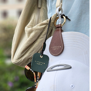 A close-up of Layover and Co. leather accessories, including a brown leather hat holder securing a white cap and a green leather sunglasses holder with gold lettering. Both are attached to a beige travel bag, showcasing their stylish and functional design for travelers. A pair of designer sunglasses hangs from the holder, adding to the aesthetic.