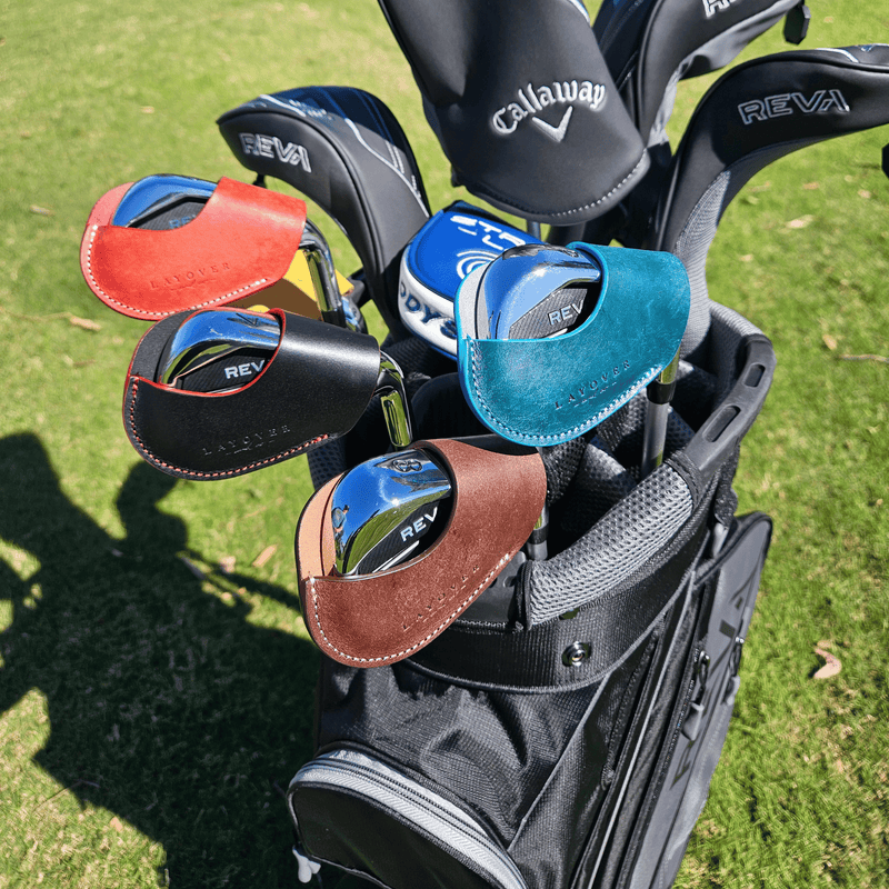 Golf clubs in a bag featuring Layover and Co. leather iron covers in red, black, cyan blue and brown in front of a golf course background