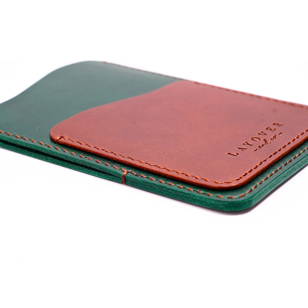 A handcrafted leather wallet featuring a deep green base and a rich brown front pocket, stitched with matching brown thread. The minimalist design is embossed with the "Layover and Co." logo, showcasing fine craftsmanship and slim functionality.