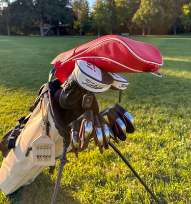 A golf bag on a grassy golf course with a red headcover, black gloves, and several metal golf clubs resting on top. The clubs show the Titleist logo.