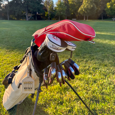A golf bag on a grassy golf course with a red headcover, black gloves, and several metal golf clubs resting on top. The clubs show the Titleist logo.