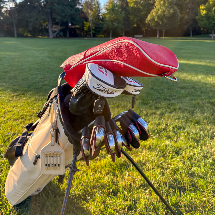 A golf bag on a grassy golf course with a red headcover, black gloves, and several metal golf clubs resting on top. The clubs show the Titleist logo.